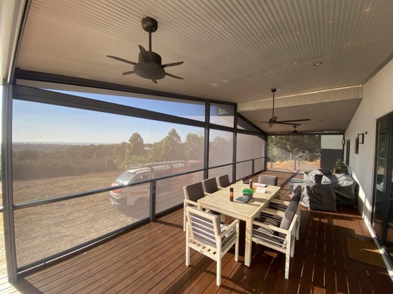 Covered alfresco with a long dining table and ceiling fan, mesh blind lowered against a distant view
