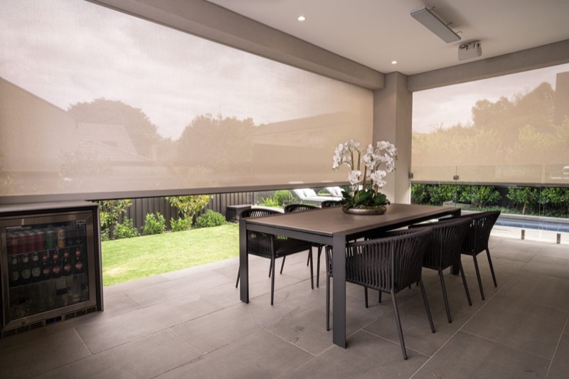 Alfresco dining setting with a ceiling fan, mesh blind lowered, lawn and trees visible through the weave