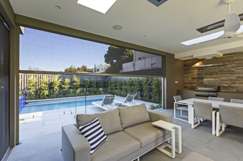 Alfresco lounge and dining space with a mesh blind lowered, looking out to a pool and garden