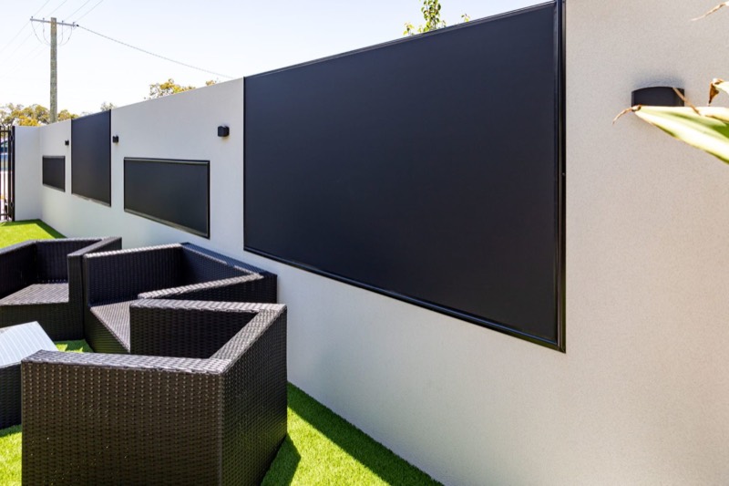 Black outdoor blind lowered against a white rendered wall with outdoor lounge seating on lawn