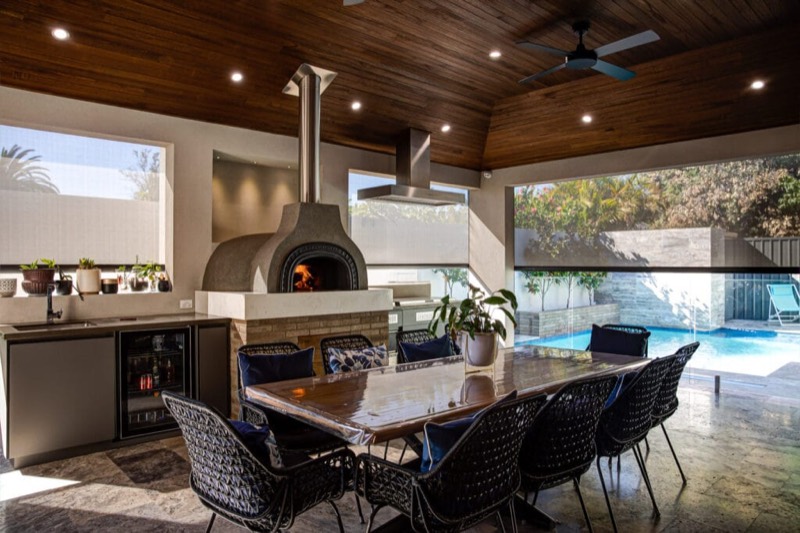 Outdoor kitchen with a pizza oven and rangehood under a timber-lined alfresco ceiling, pool beyond